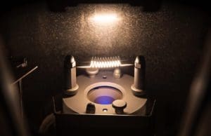 alt = An ion source from an Angstrom Engineering system. It is illuminated by a glowing filament in the center, it also has metal components. This emphasizes the detail that is essential for processes by Angstrom Engineering and the precision of their engineers.