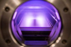 alt = close up of linear sputtering system, a metallic chamber with a sputter target. It is illuminated with a purple light. This demonstrates the precise, clean and controlled environment that Angstrom Engineering manufactures for applications.