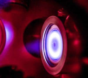 alt = close up view of a facing target sputter system. It is in operation and emitting a bright blue light. The background is illuminated by red light showing the high-energy plasma. It suggests a precise and high-technology environment created by Angstrom Engineering