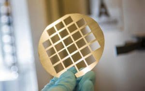 alt = close up view of a circular shadow mask. It is held by a blue gloved hand and has a grid of square cut outs. The precise design and clean edges shows the thorough job that Angstrom Engineering does.