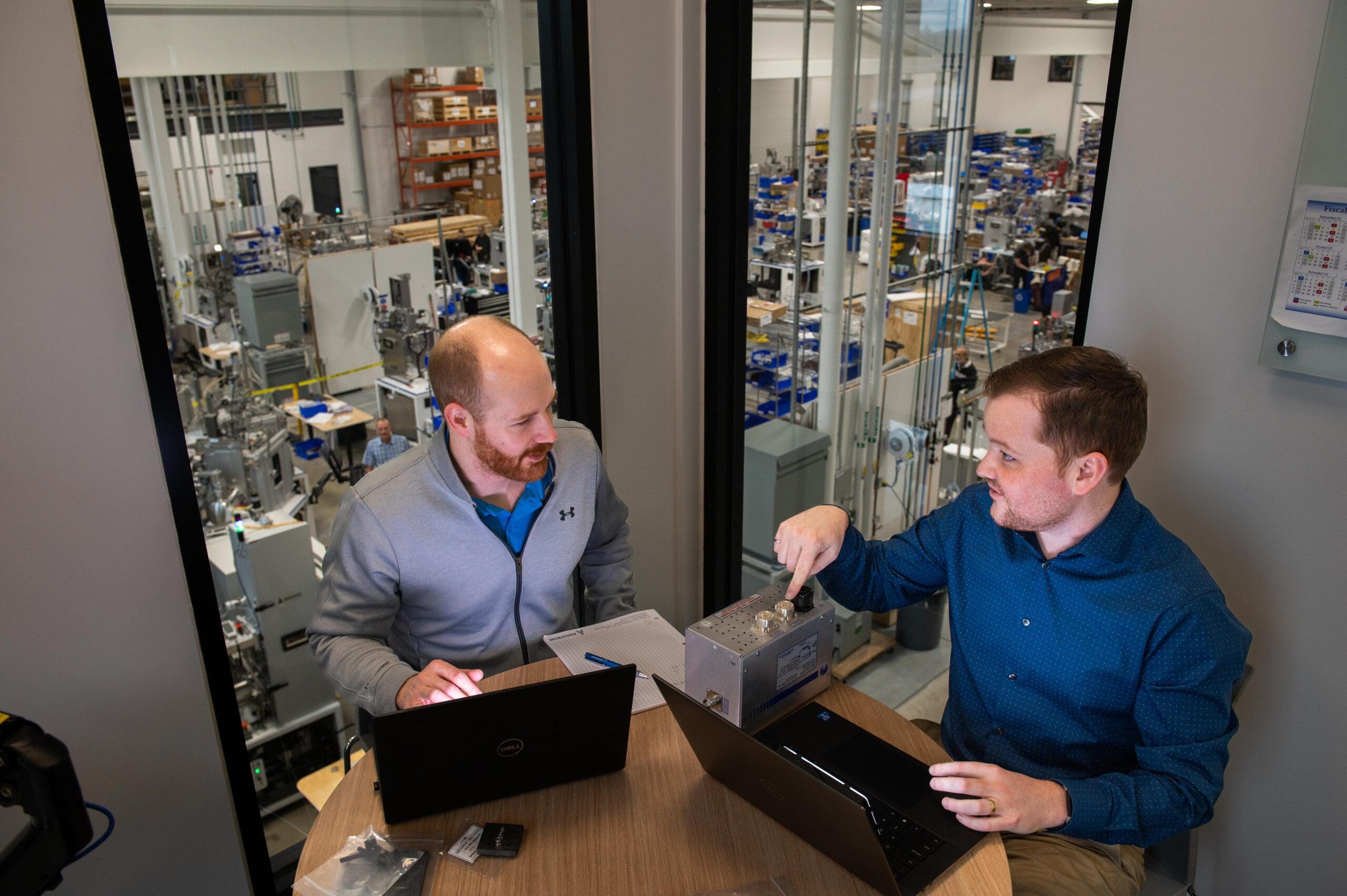 alt = Two male individuals are sitting at a round table in an office setting, both using laptops. They are in a discussion, with one person pointing to a metal component on the table. Large windows behind them have a view of a manufacturing facility with machinery and workstations. The setting shows the collaboration that plays into the work done at Angstrom Engineering and the creation of thin film deposition systems.