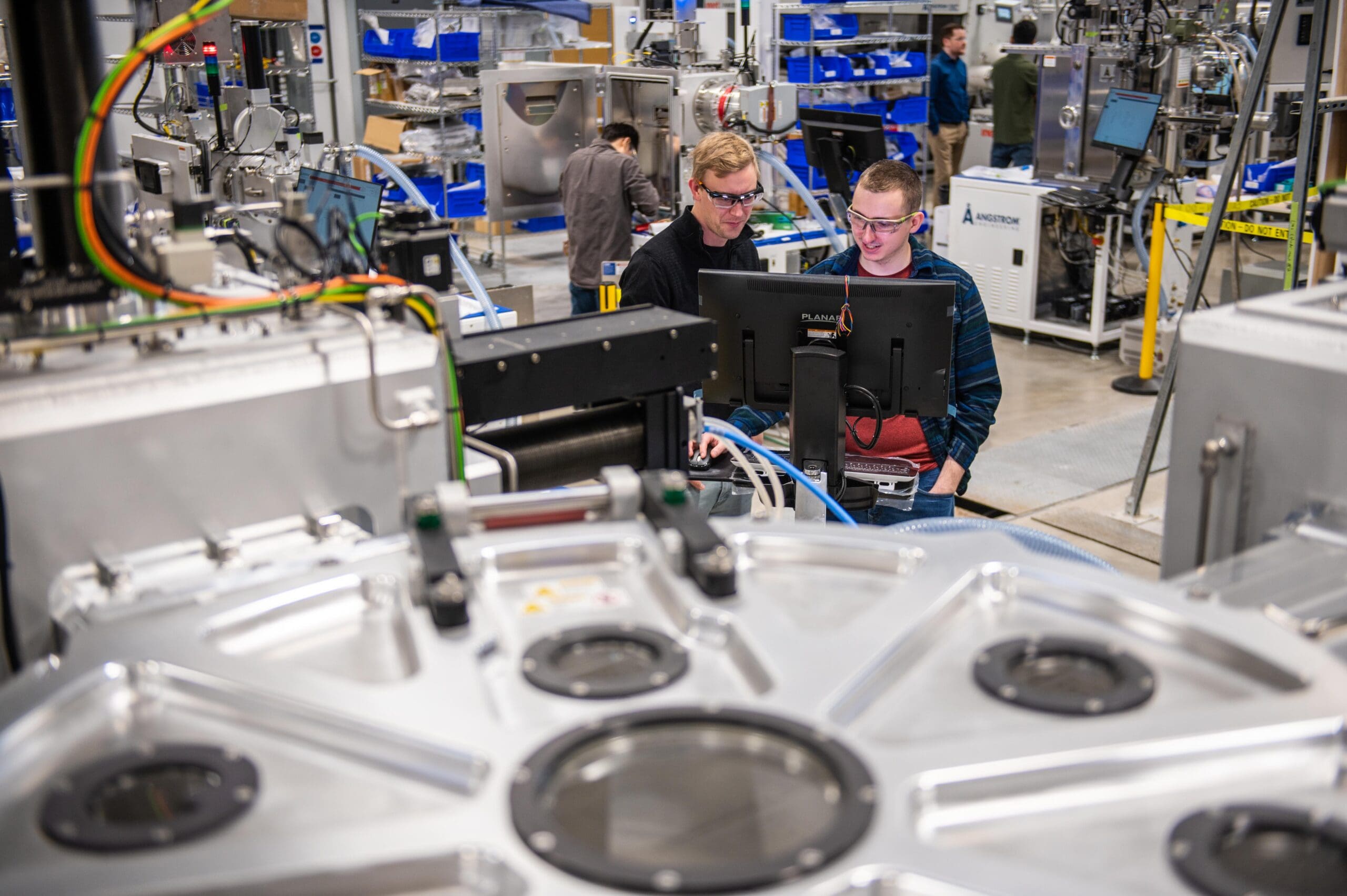 alt = Two scientists standing and working together at a digital monitor within a high-tech facility. They are surrounded by machinery , including a large metallic circular platform. Both individuals are wearing safety glasses, and the background has multiple pieces of machinery, storage bins, and others working, showing an advanced engineering and manufacturing environment.