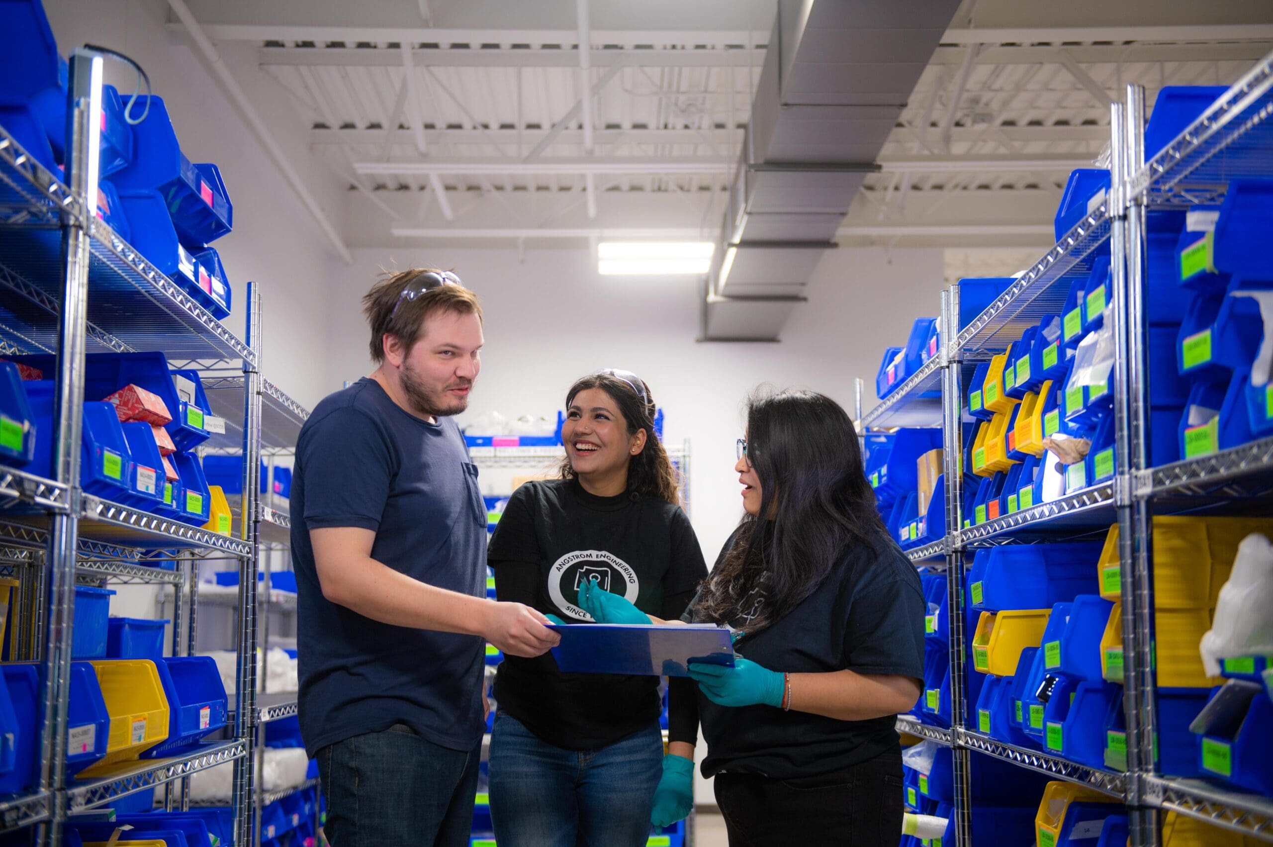 alt = Three individuals, two females and one male are standing between shelves filled with organized blue and yellow storage bins. The individuals are casually dressed in dark colours, with one person wearing safety glasses and gloves. They appear to be engaged in a friendly conversation, with one person holding a clipboard. The setting is a well-lit storage or inventory area in a professional environment symbolising teamwork’s importance at Angstrom Engineering.