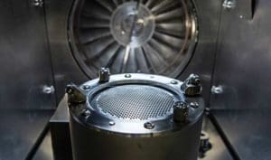 alt = close up of an ion milling source for quantum technology. It has a cylindrical component surrounded by bolts as well as other parts. There is a large fan in the background. It highlights the complexity of systems used for quantum processes.