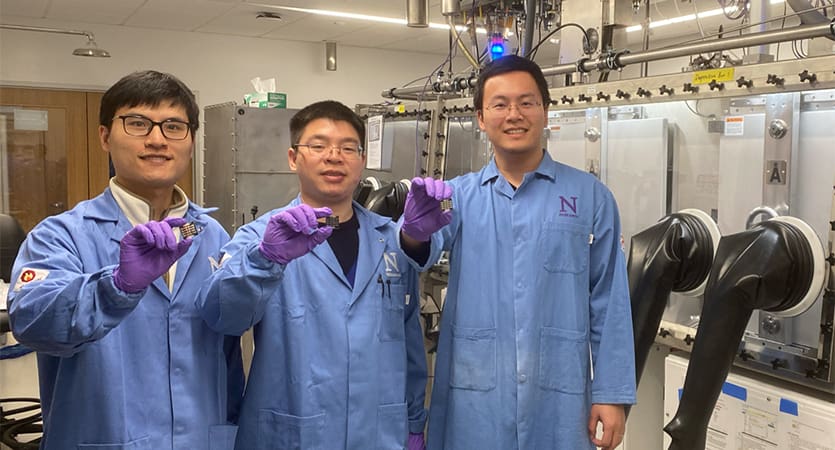 alt = Angstrom Engineering's partners at Northwestern University are seen, three male individuals are wearing blue lab coats and are each holding up a sample of material pose to celebrate setting a new world record for power conversion efficiency (PCE) of single-junction perovskite solar cells (PSCs).